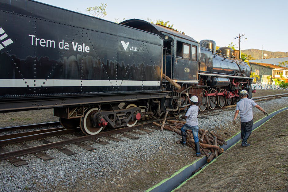A vintage black steam locomotive with red and white detailing is positioned on railway tracks outdoors, associated with the Tremo da Vale rail service. Two workers wearing white safety helmets and casual clothing are involved in a loading or unloading process; one appears to be handling wooden logs or packing materials placed on the ground, while the other is walking along the side of the train near the open side door or carriage. The scene takes place in a residential area, with houses and trees visible in the background under a clear sky, suggesting daylight conditions. The surrounding environment includes gravel on the ground near the tracks and a small paved or concrete path alongside, indicating a designated loading area. This setup may be part of logistics related to house removals or furniture transport, with the workers preparing or managing items for a home relocation, supported by Man and Van Maida Vale’s professional moving services.