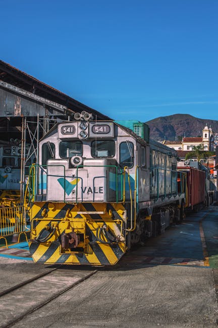 A freight train locomotive, painted in silver with a green and blue logo, is positioned on railway tracks at a station platform during daytime. The front of the locomotive features yellow and black safety markings, while the side displays several large windows and doors. Behind the locomotive, freight cars are visible, some with visible rust and wear. The platform area shows a concrete surface with metal railings and is adjacent to a building with a white facade, a small bell tower, and a red-tiled roof in the background. The scene is set outdoors with a clear blue sky and distant mountains, reflecting an industrial setting suitable for transportation and house removals involving freight or equipment transfer, as part of a professional removals service such as Man and Van Maida Vale.