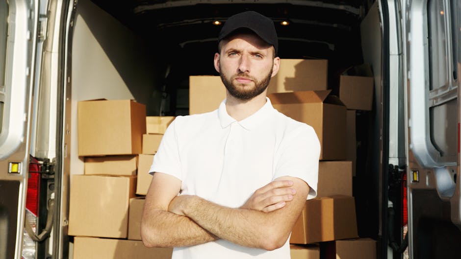 A male mover wearing a white polo shirt and a black cap stands inside an open moving van, with his arms crossed, facing the camera with a serious expression. The van's interior is filled with neatly stacked cardboard boxes of various sizes, some wrapped with plastic film, positioned on the floor and shelves. The background includes a partially visible building exterior, indicating the loading or unloading area for home removal or furniture transport. The scene is well-lit, capturing the details of the packing and loading process involved in house removals managed by Man and Van Maida Vale, highlighting the importance of proper organization during furniture transport and relocation services.