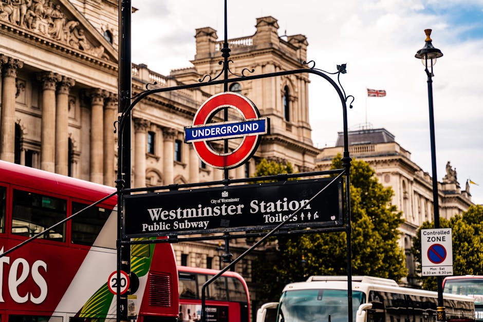 Close-up view of a black metal signpost with a red and blue London Underground roundel for Westminster Station, indicating the entrance to the public subway. The sign attached beneath displays the station name, 'Westminster Station,' along with details about toilets and staff. The background features a historic neoclassical building with ornate stone architecture, large windows, and decorative columns, set against a partly cloudy sky. There is a London bus with red and white livery passing nearby, along with street lighting and a controlled zone traffic sign visible on the right side. During a house relocation or furniture transport, such signage and urban surroundings are typical of central London streets, providing context for moving logistics in areas managing traffic and pedestrian access, relevant for professional removals services like those offered by Man and Van Maida Vale.
