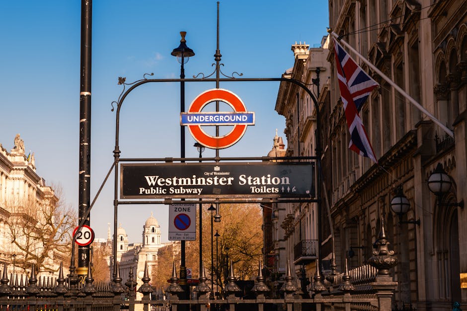 Close-up view of the entrance sign to Westminster Underground Station in Maida Vale, London, showing the classic red and blue circular underground logo and a black rectangular sign with white text indicating 'Westminster Station' along with directions for public subway and toilets. The sign is mounted on a black metal structure attached to a lamppost, with a Union Jack flag visible on the right side. In the background, ornate historic buildings with decorative facades and a clear blue sky can be seen, with trees showing autumn foliage. This scene reflects a typical urban environment where home relocation and furniture transport services by Man and Van Maida Vale may take place, especially considering city traffic regulations and the importance of logistical planning for removals in the area.
