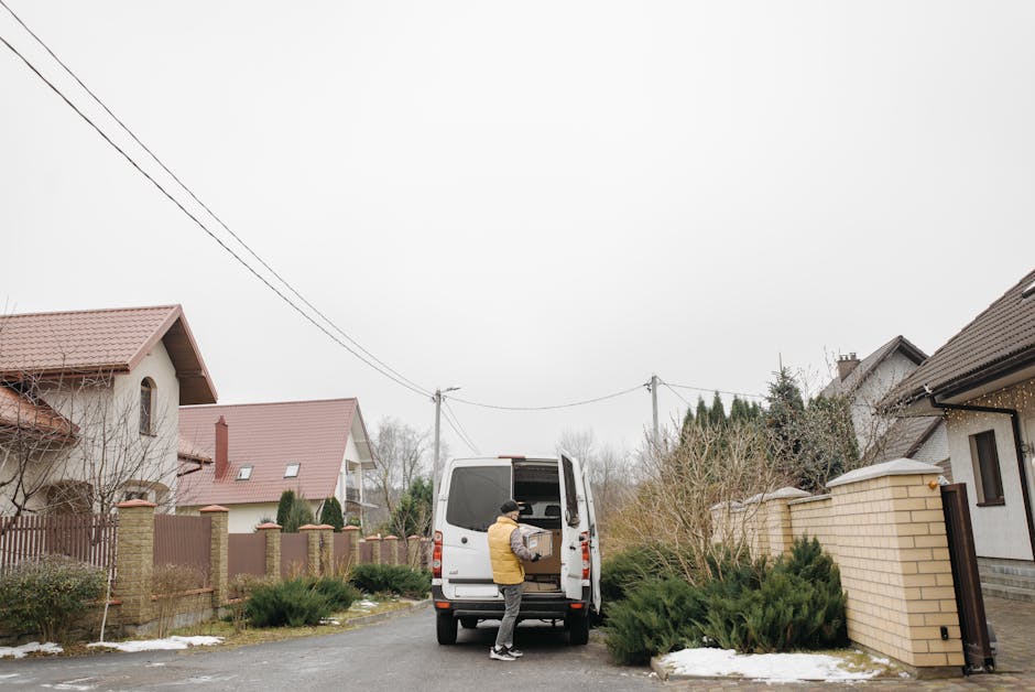A man wearing a yellow jacket and black trousers loading a cardboard box into the rear of a white van parked on a residential street. The van, part of a home removal service by Man and Van Maida Vale, is positioned near the pavement adjacent to a suburban property with a brick wall and garden bushes. The street is lined with typical houses featuring pitched roofs, some with small skylights, and garden fences. Overhead, electrical wires stretch between wooden utility poles against an overcast sky. Patches of snow are visible on the ground and on some shrubbery, indicating winter weather. The scene captures the loading process during a house relocation, with the van's rear doors open, and the man beginning to carry or load packing boxes, aligning with furniture transport and packing and moving operations typical of house removals in Maida Vale.