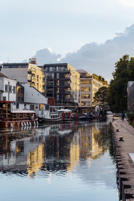 Riverfront view in Little Venice Maida Vale showing modern residential buildings with large windows and balconies, situated along a canal with calm water reflecting the structures above. On the right, a paved walkway runs parallel to the water, lined with small bollards and greenery, with a few pedestrians visible. On the left side, a houseboat with outdoor seating and a small floating dock are positioned near the water's edge. The scene is illuminated by natural daylight, with partly cloudy skies overhead. This image depicts a peaceful urban setting suitable for house removals and furniture transport services offered by Man and Van Maida Vale, focusing on the logistics of home relocation in canal-side properties.