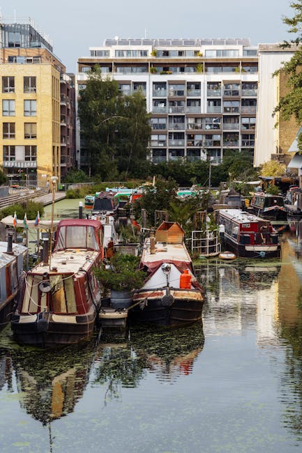 A canal lined with several narrowboats painted in shades of blue, purple, and black floats on calm water, with trees displaying green, orange, and red foliage along the banks. The sky above is partly cloudy with patches of blue visible. On the left, a paved walkway runs parallel to the canal, and the boats are moored close to the edge, some with visible covers and rooftop equipment. The scene depicts a peaceful, scenic setting typical of house removals and relocation services near waterways, with no visible activity but suggesting a quiet environment suitable for moving preparations or storage near canal side homes. The overall image supports the context of house removals in Maida Vale, especially in locations close to waterways like Little Venice, as managed by Man and Van Maida Vale.