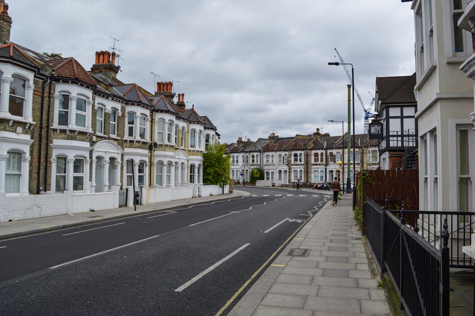 A residential street in Maida Vale W9 lined with traditional Victorian-style terraced houses featuring white facades, bay windows, and red-tiled roofs under a cloudy sky. On the right side, there is a sidewalk with a black metal railing, and a person walking away from the camera, carrying a cardboard box as part of a home relocation process. Visible parked cars are along the curb, and a narrow empty road with road markings extends ahead. Overhead, there are streetlights and a construction crane in the distance, indicating ongoing building work. The scene illustrates a quiet, urban neighbourhood suitable for house removals and furniture transport, with [MAN AND VAN MAIDA VALE] often servicing such property moves in the area.
