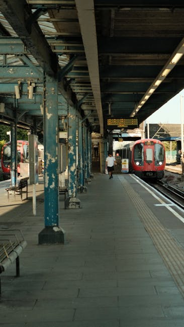 The image depicts a train station platform with an overhanging roof supported by weathered blue metal columns. On the platform, there is a person dressed in casual clothing walking away from the camera, towards the arriving train. The train, which is red in colour, is stationary on the track adjacent to the platform, with its doors closed. The platform surface is made of concrete with tactile paving near the edge for safety, and there are empty metal benches along the side. Overhead lighting is visible, mounted underneath the platform roof, and a digital information display is hanging from the ceiling showing train schedules. Natural light filters in from the station's open sides, creating a well-lit environment suitable for passenger movement, aligning with transport and local travel services, such as those offered by Man and Van Maida Vale for home relocation and furniture transport purposes.