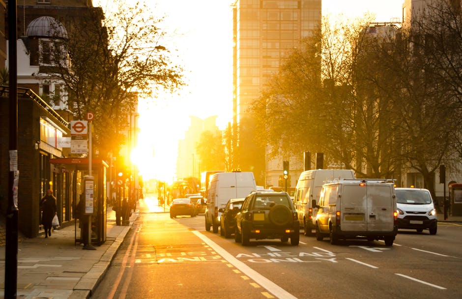 A busy urban street scene during sunset in Maida Vale, London, with a row of white and dark-colored vans parked along a designated loading zone, which is marked with white lines on the asphalt. Several vehicles are parked closely together, while others are in motion or waiting at a traffic light further down the road. On the left, a bus stop shelter with timetable posters is visible, with a few pedestrians walking along the sidewalk, some carrying bags. Tall buildings and modern skyscrapers rise on both sides of the street, illuminated by warm, golden sunlight filtering through the trees lining the pavement. The trees, with sparse autumn foliage, cast shadows on the sidewalk, and the overall scene suggests a busy area suitable for home relocation or furniture transport operations carried out by [COMPANY_NAME], supporting efficient house removals and moving services in Maida Vale.