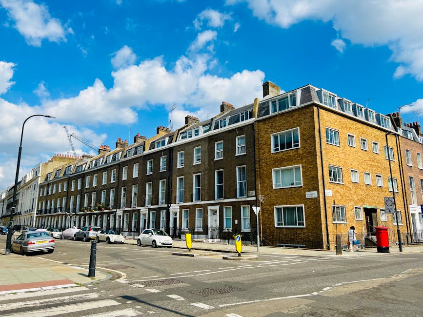 A multi-storey residential building with a brick facade and white-framed windows, situated on a busy urban street in Maida Vale. The building features a sloping roof with multiple dormer windows, and balconies on the lower floors. Parked cars line the street in front, including a white van that is likely used for house removals or furniture transport. The sidewalk is populated with pedestrians, and a red post box is visible near the corner. Overhead, a lamppost stands alongside a bright blue sky with scattered white clouds. The scene reflects a typical setting for home relocation services, where furniture and removal equipment may be prepared for loading or unloading, aligned with the services provided by Man and Van Maida Vale.