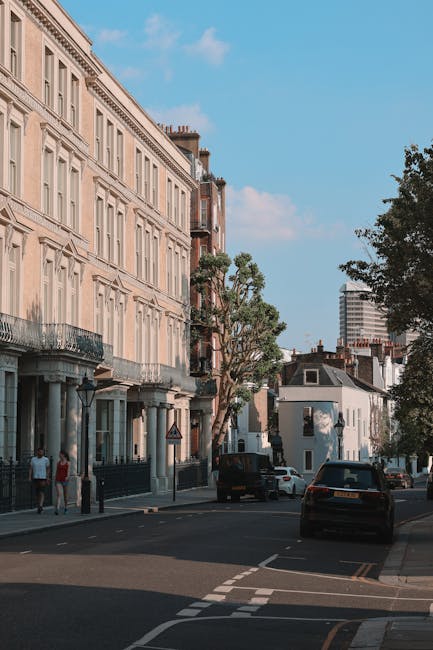 The image depicts a typical residential street scene in Maida Vale with a clear blue sky overhead. On the left side, there is a row of multi-storey Victorian-style terraced buildings with ornate facades, large windows, and small balconies with wrought iron railings. The street is lined with trees, one of which has a large, pruned canopy, while others have dense green foliage. Parked along the curb are several cars, including a black van parked directly in front of the homes, likely used for furniture transport during a home relocation. The street has a paved sidewalk where a few pedestrians are walking, and a man in casual clothing is seen near a corner street sign. The environment suggests a peaceful neighbourhood suitable for house removals and professional moving services. The presence of the van and the condition of the surroundings indicate that a moving company, such as Man and Van Maida Vale, might be involved in packing and loading furniture and boxes for a house move, with items possibly inside the van or ready to be loaded onto it for transport.